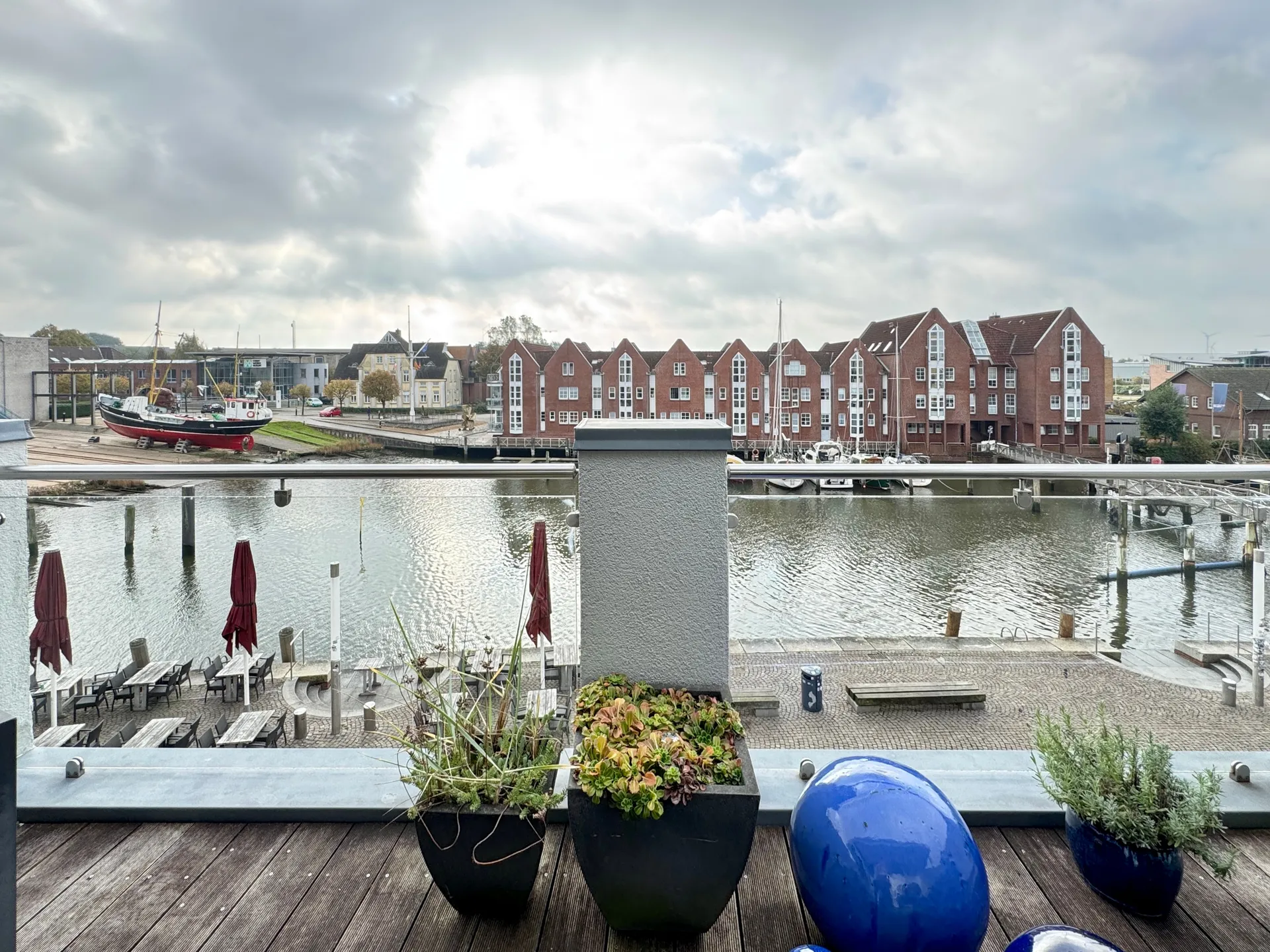 Blick von der Dachterrasse über den Husumer Binnenhafen auf die Giebelhäuser aus rotem Backstein, links ein Kutter an Land, im Vordergrund Pflanzkübel auf dem Holzboden der Terrasse