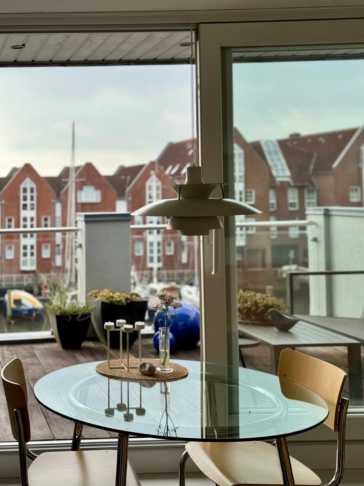Runder Glastisch mit zwei Stühlen am großen Fenster, dahinter die Dachterrasse und die Giebelhäuser am Hafen